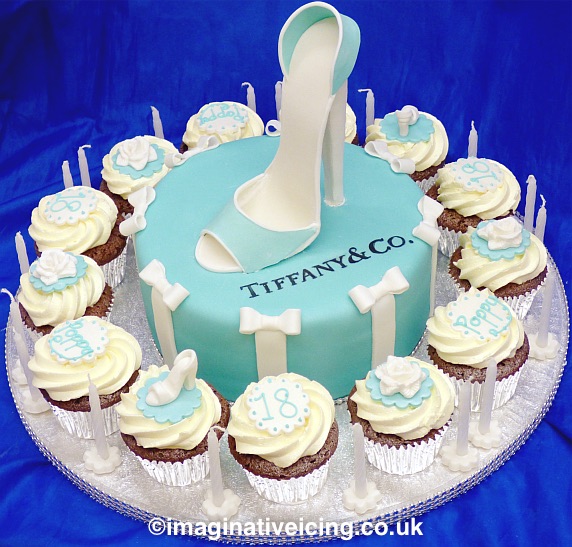 High heeled shoe made from icing on top of a round cake iced in bluey green turquoise tiffany blue. Icing bows and icing ribbon up sides. Cupcakes in silver cake cases with swirl of buttercream and icing decorations, piped age and name. Round Silver Cake board with Diamonte ribbon trim and 18 birthday candles. Designer logo painted on cake.