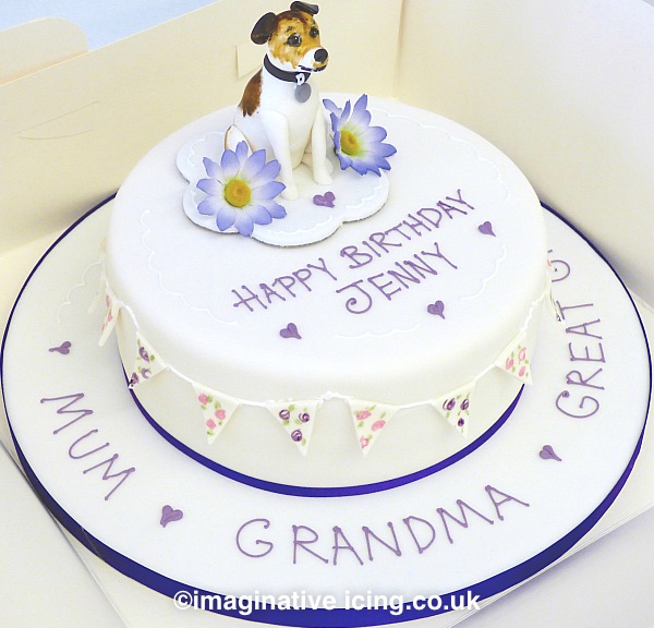 Great Granny's Jack Russel Birthday Cake - Birthday cake iced in white with edible flowery bunting round sides, Icing model of a jack Russel on the top with flowers either side. Inscription piped on top of cake and on cake board in lilac icing