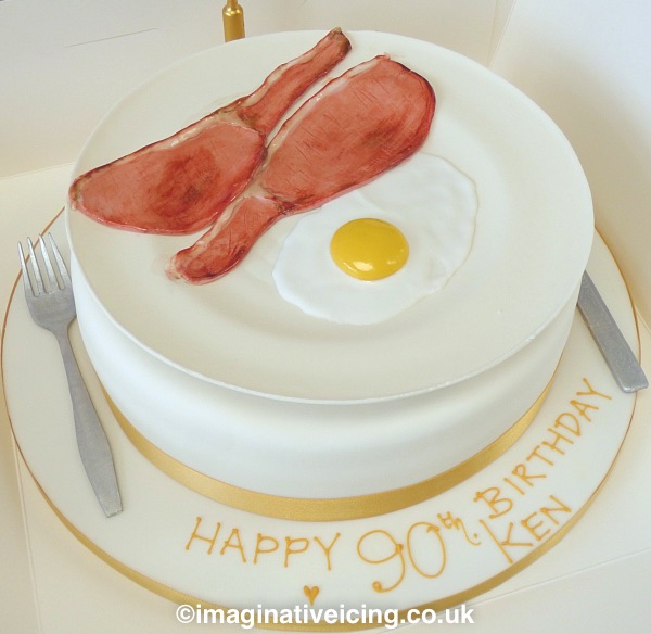Bacon and Egg on a plate 90th Birthday Cake. Every thing you see is made from icing even the knife and fork and the plate. Placed on a round cake base iced in white. trimmed with golden ribbon and on a iced cake board. Inscription piped in golden royal icing